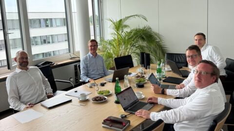 Fünf Männer sitzen an einem Besprechungstisch und schauen in die Kamera.