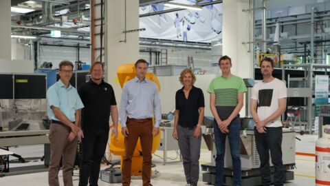 Fünf Männer und eine Frau stehen nebenaindner in einer Produktionshalle und schauen in die Kamera.