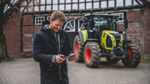 Ein Mann steht vor einem Trecker und blickt auf sein mobiles Endgerät