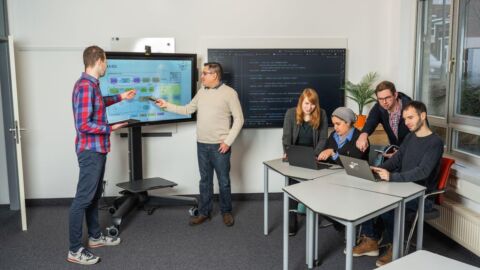 Mehrere Personen in einem Raum arbeiten an einem Bildschirm und an einem Tisch mit Laptops