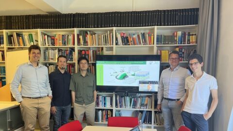 Fünf Männer stehen in einem Meetingraum, im Hintergrund ist eine Regal mit Büchern zu sehen, davor ein Monitor mit einer Grafik.