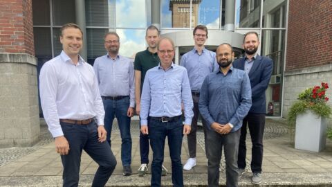 Sieben Männer stehen vor einem Bürogebäude und lächeln in die Kamera.