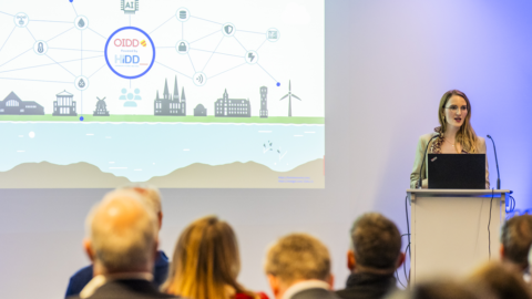 Frau steht vor einem Rednerpult und schaut ins Publikum, das man von hinten sieht. An der Wand hinter Frau ist eine Leinwand mit einer Präsentation zu sehen.